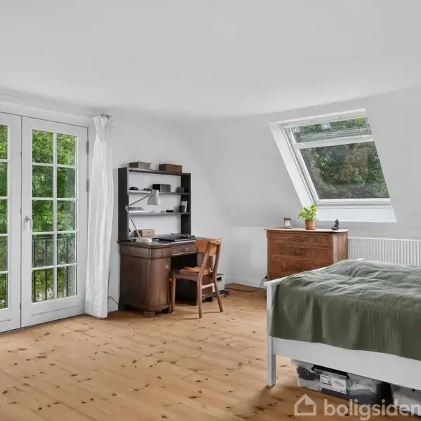 Landsbymiljø omgivet af grønne træer, inkluderer huse med røde og grå tage i forskellige størrelser. Beliggende ved veje med rabatter. Malerisk landskab i baggrunden.