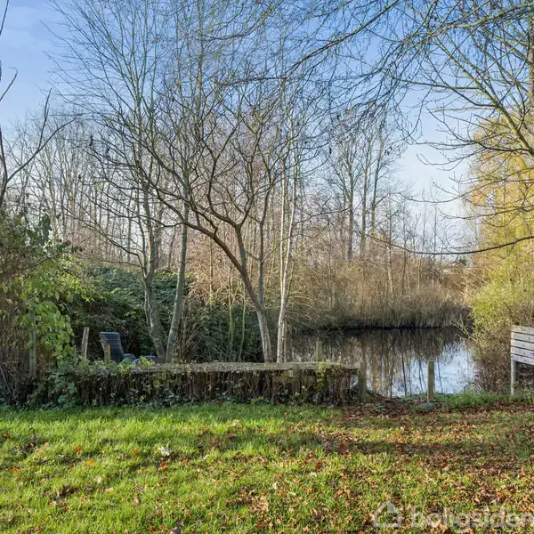En have med tæppe af græs omkranser et lille, stille vandløb omkranset af træer og buske i vinterdragt, med to stole ved en træterrasse og en lille skænk.