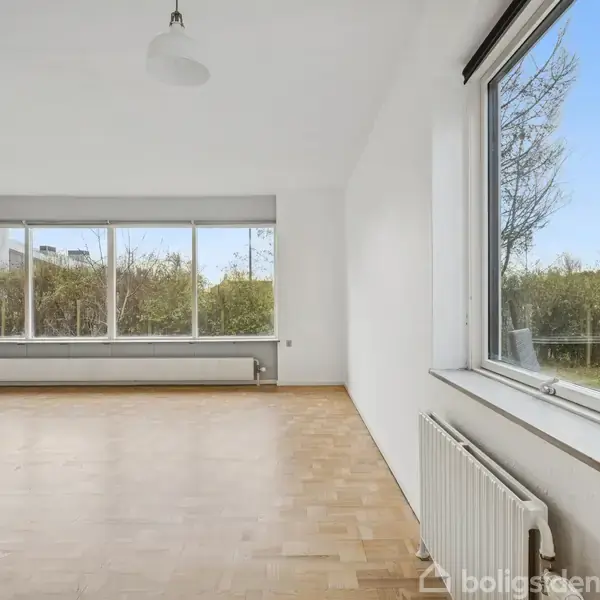 Et lyst, tomt rum med trægulv og store vinduer. Udsigt til en hæk og blå himmel udenfor. Radiatorer under vinduerne, og en pendellampe hænger fra loftet.