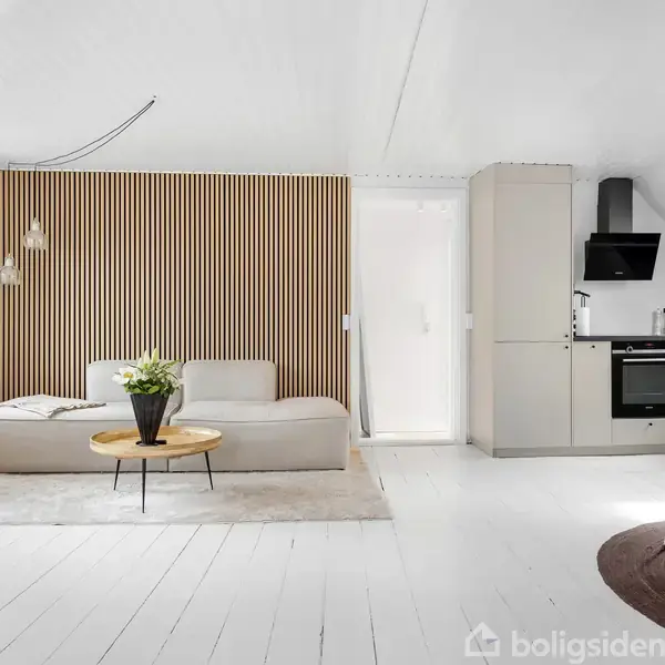 Sofa står foran en træpanelvæg med hængende lamper. Lille rundt bord med blomster. Lyse køkkenskabe og komfur i baggrunden. Rummet er lyst med hvidmalede gulve.