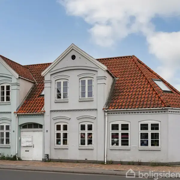 Hvidt hus med rødt tegltag, står langs en vej i en by. Det har tre sektioner, hver med symmetriske vinduer og indgangsdør.