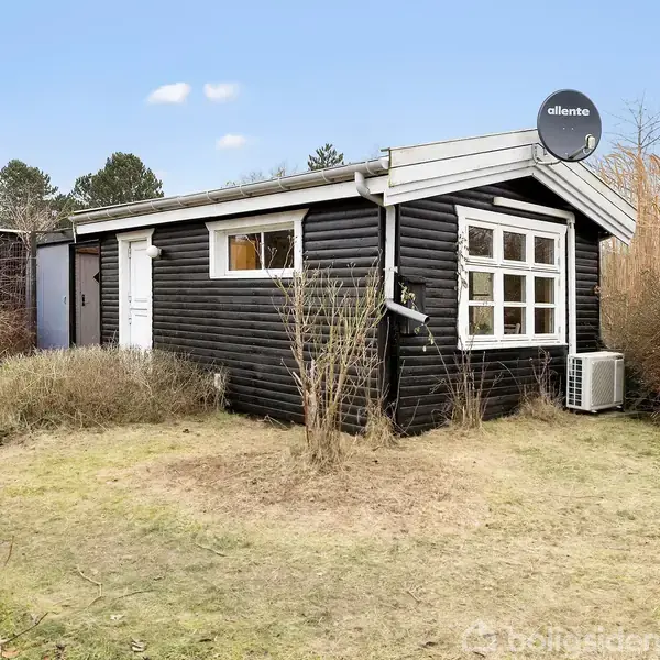Sort træhus står stille på en græsplæne omgivet af buske og træer, med en parabolantenne, "allente," på taget og en airconditionenhed ved siden af vinduet.