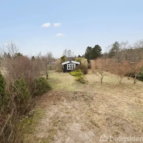 Et lille, sort træhus med hvide vinduer står i en stor, åben have omkranset af træer og buske, under en klar blå himmel. En bil er parkeret til venstre.