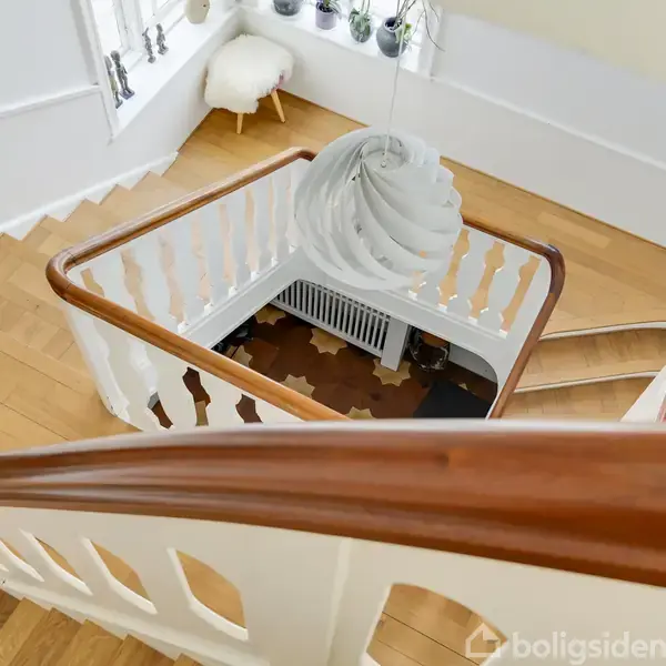 Trappe med hvidt gelænder og træhåndliste drejer nedad i et lyst rum. Over trappen hænger en moderne, spiralformet lampe. En stol står ved et vindue med planter.
