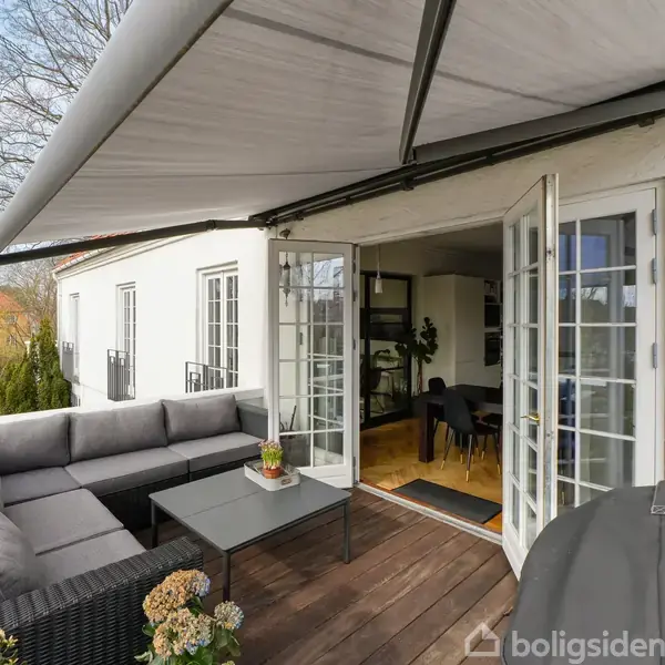 En moderne terrasse med grå hjørnesofa og bord. Åben dobbeltdør fører til indendørs spisestue. Terrassemarkise skaber skygge, mens grønne træer omgiver området.