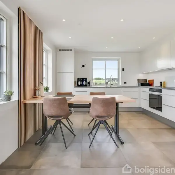 Et moderne køkken med en træbordplade og fire stole står i centrum. Indbyggede hvide skabe og apparater omkranser rummet. Store vinduer på venstre side giver udsigt til grønne omgivelser.