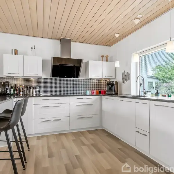 Lys køkken med hvide skabe, sort bordplade og moderne apparater, inklusive ovn og emhætte. To høje stole ved en bardisk. Træloft og stort vindue med udsigt til have.