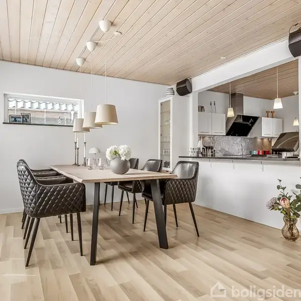 Spisebord med seks stole står i en moderne spisestue. Bordet er dekoreret med blomster og lamper. Ved siden ses et åbent køkken, træloft og parketgulv.