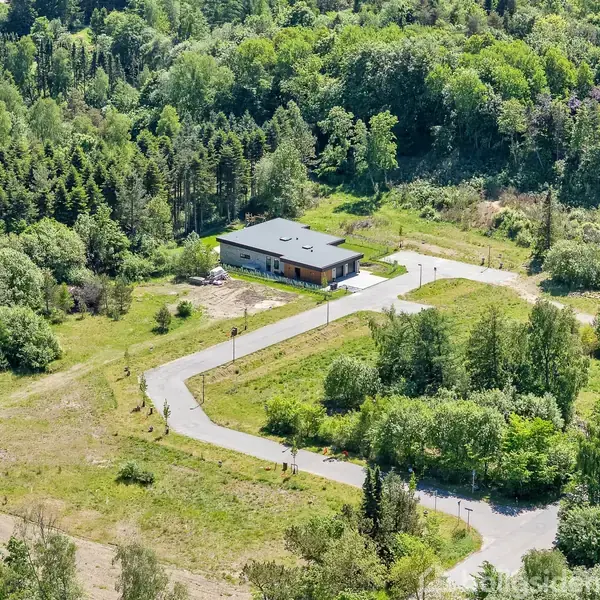 En moderne, fladtagshus beliggende midt i en stor, frodig skov. Huset er omgivet af en grusvej og åbne, grønne områder med tæt bevoksning i baggrunden.
