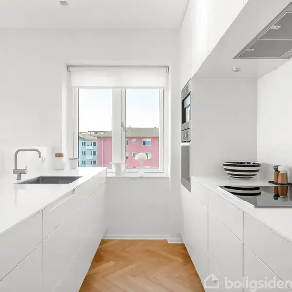 Hvidt moderne køkken med kogeplade, indbyggede ovne og vask. Stort vindue i baggrunden viser udsigt til farverige bygninger udenfor. Parquet trægulv tilføjer varme. Ingen tekst i billedet.