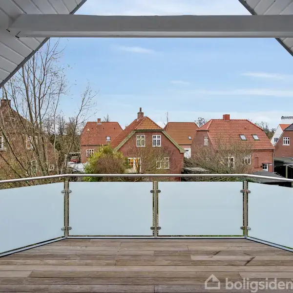 En balkon med glasrækværk og trægulv vender ud mod en række villaer med røde tage i et boligkvarter. Træer uden blade står omkring husene.