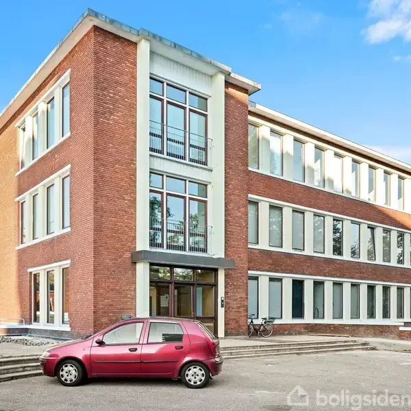 En rød bil holder parkeret foran en stor murstensbygning med mange vinduer. Bygningen har tre etager og ligger i et åbent område med en klar, blå himmel.
