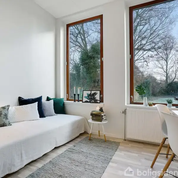 En enkel stue viser en hvid sofa med farverige puder mod en hvid væg. Store vinduer giver udsigt til træer udenfor. Et lille bord med plante, lamper og stole danner hygge.