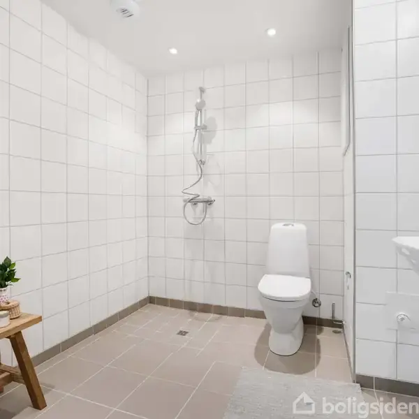 Et hvidt flisebelagt badeværelse med toilet og bruser til venstre. En håndvask er til højre. En træbænk med kurve og plante står mod venstre væg.