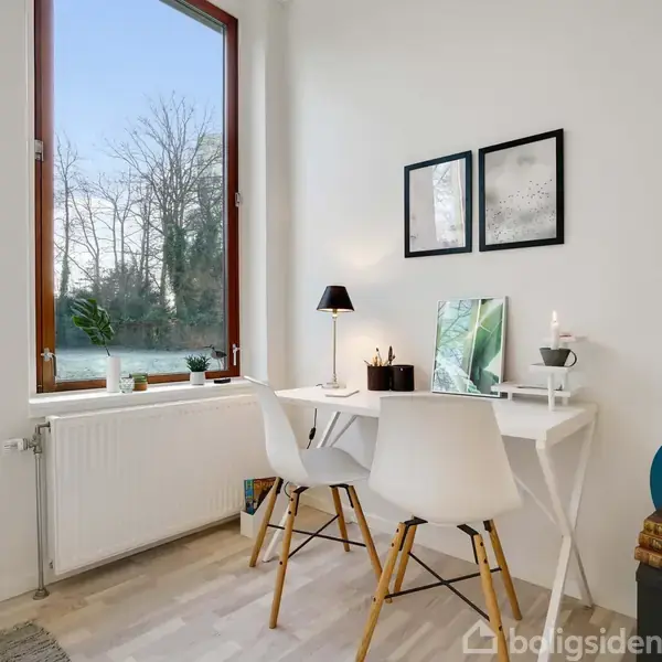 Et hvidt skrivebord med to hvide stole står ved et stort vindue. På bordet er en lampe, planter og dekorationer. Rummet har lyse vægge og et trægulv, med bøger stablet ved siden af.