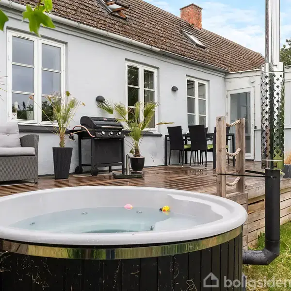 Udendørs spabad står fyldt med vand og plastikænder. Ved siden af er en træterrasse med grill, sofagruppe og spisebord foran et hvidt hus med flere vinduer og en græsplæne.
