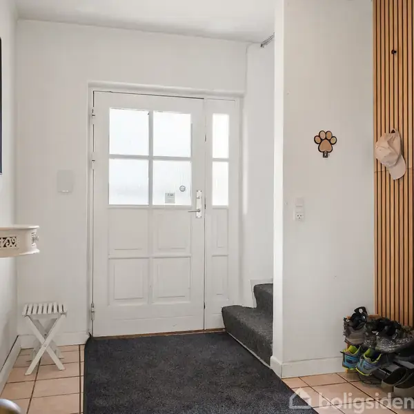 En entré med en hvid dør i midten, flankeret af et vindue. Et lille, hvidt bord hænger til venstre. Sko og en jakke hænger på vægpanelerne til højre. Tekst: "Welcome to our home."