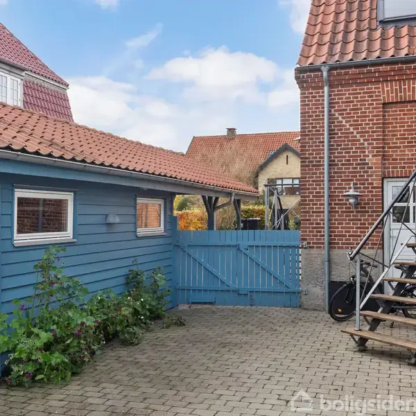 En gårdsplads med brosten, omkranset af en blå træbygning og en murstensbygning. En trappe med cykel ved siden af leder op til en dør. Blå port i baggrunden.