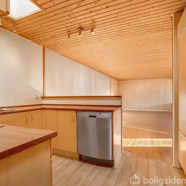 Et køkken med lyse træskabe, indbygget ovn og en opvaskemaskine. Rummet har træloft og gulv, og der er naturligt lysindfald fra en loftsåbning. Ingen tekst er til stede.