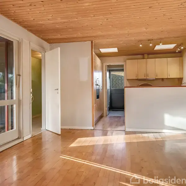 Stue med trægulv og træloft, sollys strømmer ind gennem store vinduer. Åben dør til køkken med lyse skabe. Rummet er lyst og rummeligt uden møbler.