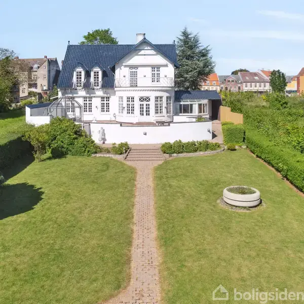 Et hvidt hus med sort tag står foran en stor, velholdt have med græs. En murstenssti fører op til huset, omgivet af buske og trimmet hæk.