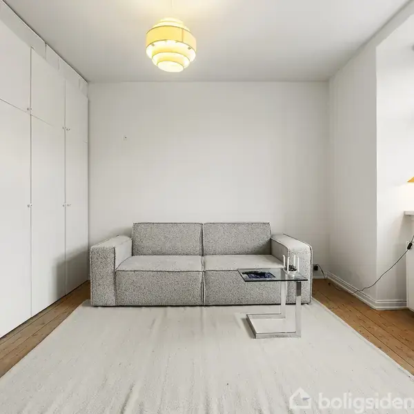 Sofa står stille på lys tæppe i en minimalistisk stue. Ved siden af er et lille bord med magasiner. Garderobeskabe dækker venstre væg, og et vindue slipper lys ind.
