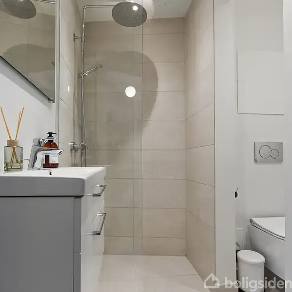 Brusekabine med glasdør står i et smalt, lyst badeværelse. Håndvask med sæbedispenser og reed diffuser er ved siden af. Toilettet er til højre for bruseområdet.