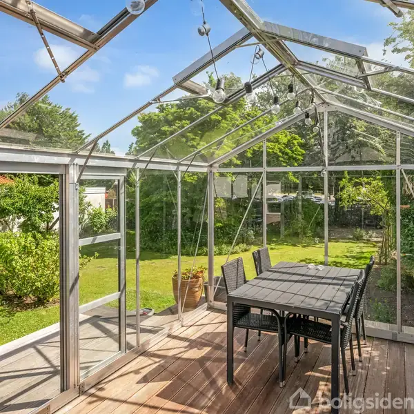 Et bord med stole står i en glaspavillon i en frodig have. Pavillonen har klare paneler, der giver udsigt til grønne træer og buske under en blå himmel.