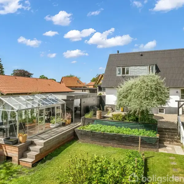 Hus med mørkt tag, placeret i en velholdt have, hvor en glaskonstruktion fungerer som drivhus til planter. Græsplæne og blomsterbede omgiver området. Solrig dag med få skyer.