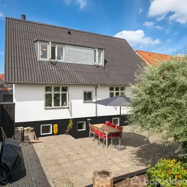 Et to-etagers hus med hvid facade og sort tag. På terrassen er der et bord med stole under en parasol. Haven er stenbelagt med træer og buske omkring.