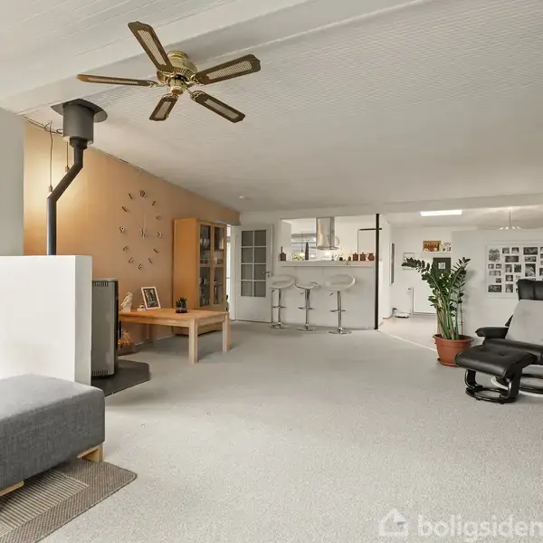 En lys stue med sofa og fodskammel, brændeovn og loftventilator. I baggrunden ses et åbent køkken med barstole og en spiseplante ved siden af en lænestol. Ingen tekst til stede.