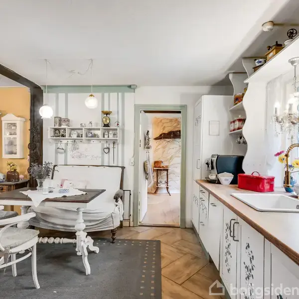 Et landligt køkken med hvide møbler står stille i et hyggeligt rum. Til venstre er der et spisebord med stole, og til højre er der et køkkenbord med vask.