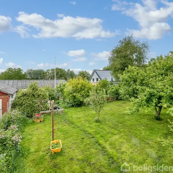 En grøn have med en gynge hængende fra en tørresnor. Til venstre ses en rød bygning, omgivet af træer og buske under en blå himmel med spredte skyer.