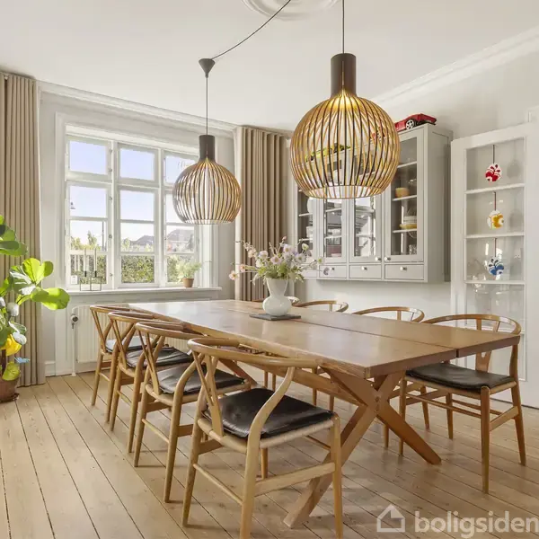 Et spisebord med seks stole står i en lys spisestue med trægulv. To moderne lamper hænger over bordet. I baggrunden ses en stor plante og et vitrineskab.