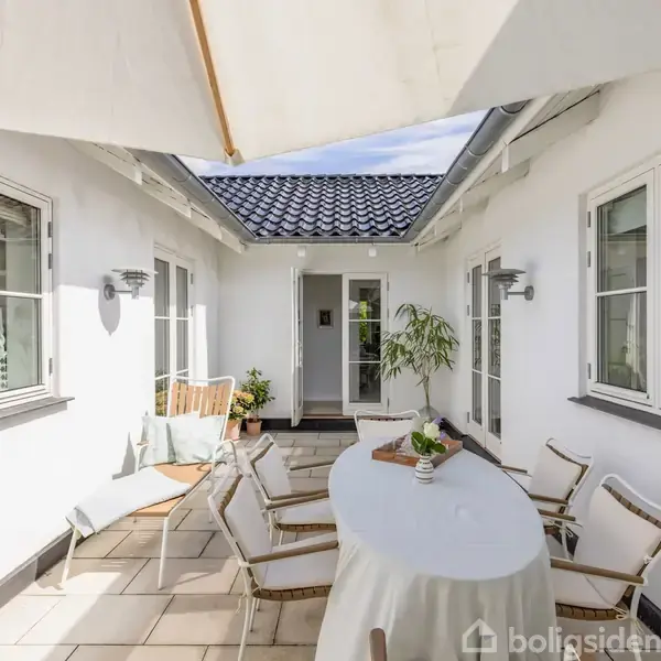 Et rundt bord med hvid dug står i centrum omgivet af stole. Bordet er pyntet med en plante. I en lys indendørs gårdhave med hvide vægge og vinduer. En parasol skaber skygge.