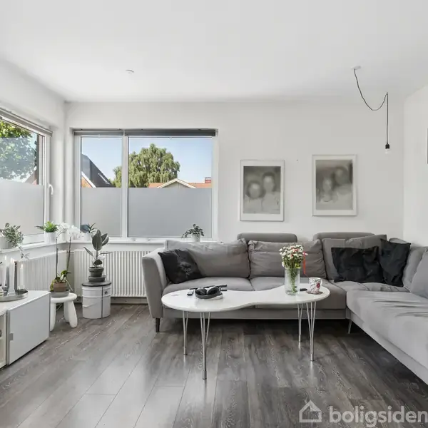 Stue med grå hjørnesofa, dekorative puder, og et sofabord med blomster. Et fjernsyn er monteret på væggen. Rummet har store vinduer, planter og lysegrå vægge med kunstværker.