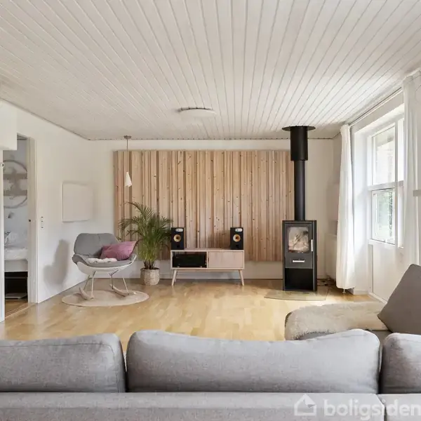 Stue med grå sofa i forgrunden. En brændeovn står nær trævæg med højttalere. Lyse vægge, trægulv, store vinduer og planter skaber hyggelig atmosfære. En stol med pude er i hjørnet.