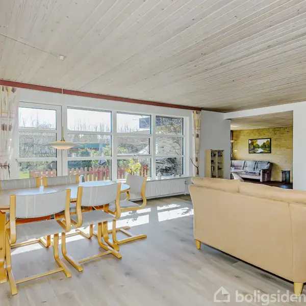 Spisebord med stole står ved store vinduer, der lukker lys ind. Sofa er placeret til højre i stuen, som er rummelig og lys.