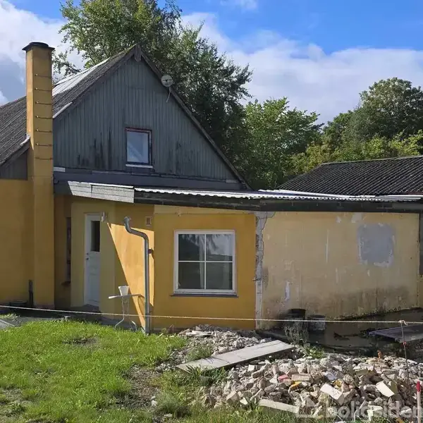 Et faldefærdigt gult hus står i en have med græs. En del af facaden er under renovering, og der er byggeaffald foran. Træer omkranser huset.