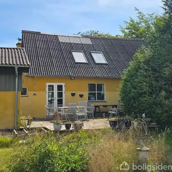 Gulmalet hus med skråt tag og ovenlysvinduer. En terrasse foran har havemøbler og potteplanter. Haven er delvist tilgroet med græs og buske omgivet af træer.