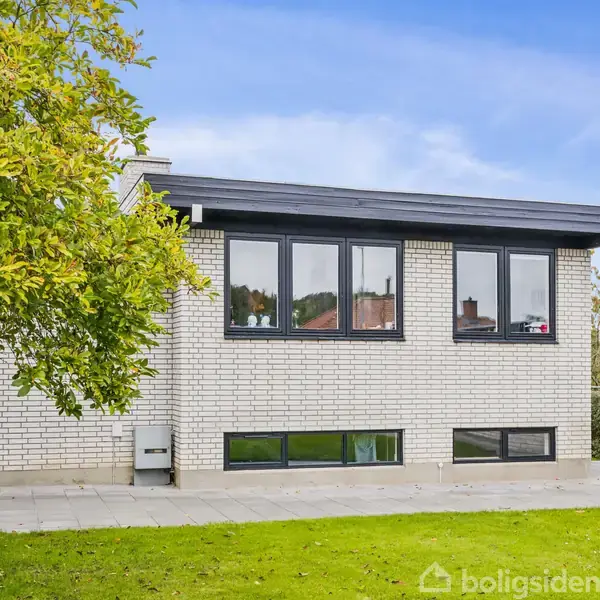 Hus med hvid murstensfacade og sorte vinduesrammer, omgivet af grøn plæne og træ, under blå himmel.