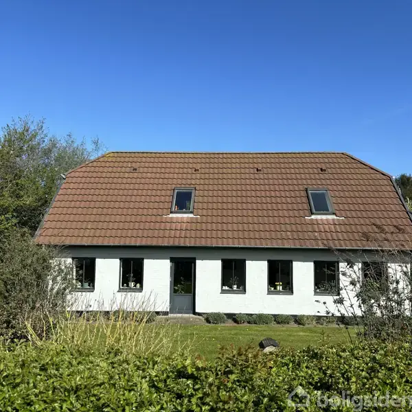 Hus med hvid facade og brunt tegltag har fem vinduer, en dør foran og er omgivet af grønne buske og træer under en klar blå himmel.