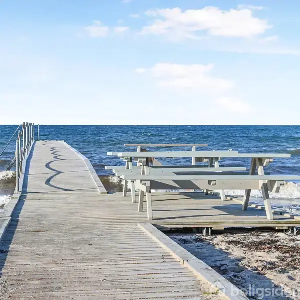 En træbro strækker sig ud over havet med bølger, der bryder ved siden af. Broens side har to picnicborde og omgivelserne er himmelblå med lette skyer.
