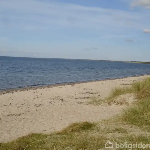 Strand langs kystlinje med klart blåt hav på højre side, bølger der let berører sandet. Grønne klitter med græsset omkranser stranden.