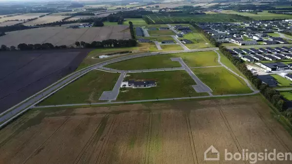 Grønt feltområde under udstykning ligger midt i en landlig landskabskontekst med marker i baggrunden og et nærliggende parcelhuskvarter.