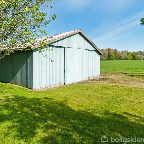 Et lyseblåt skur står stille på en grøn græsplæne med træer og en stor mark i baggrunden under en klar blå himmel.