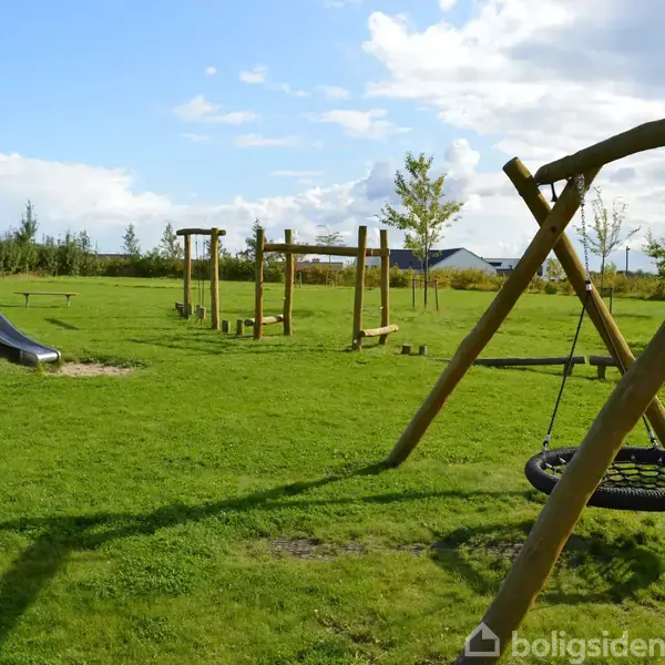 En legeplads med en gyngestativ i træ står i en grøn park. En rutsjebane og klatrestativ kan ses i baggrunden under en blå himmel med skyer.