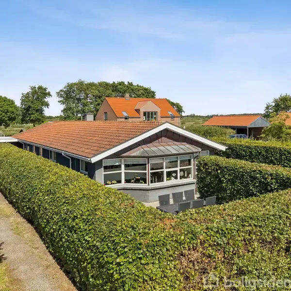 Et ensomsiddende, moderne hus med rødt tegltag og store vinduer er omgivet af klippede hække. En grusvej løber langs huset, der er placeret i et grønt boligområde.