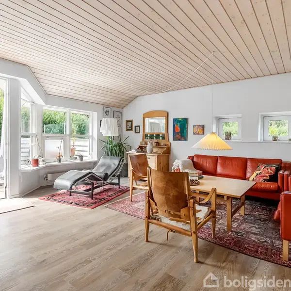 Stue med rødt lædermøblement, herunder sofa og stole, stående på tæpper. Store vinduer og dør til have. Træloft og -gulv giver rummet en lys, åben atmosfære.