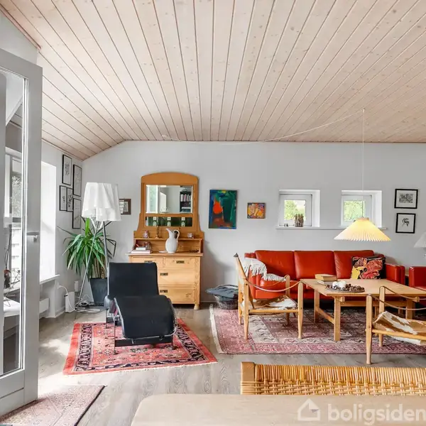Stue med afslappet atmosfære: To sorte lænestole og en rød sofa omkranser et træbord. Væglamper og indrammede billeder pryder de lyse vægge. Store vinduer giver lysindfald.